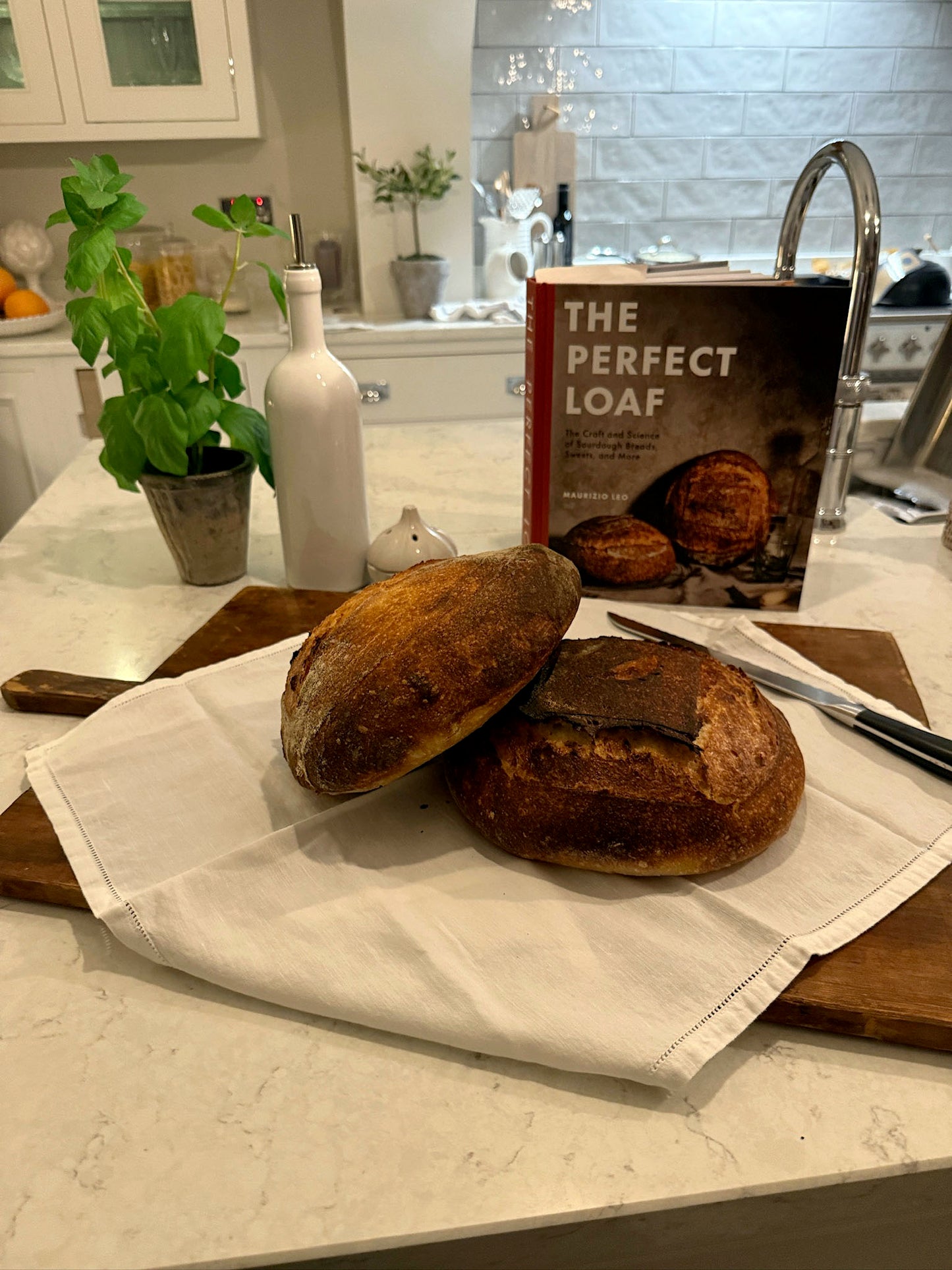 Two loaves of bread on a kitchen counter with a cookbook titled 'The Perfect Loaf'.