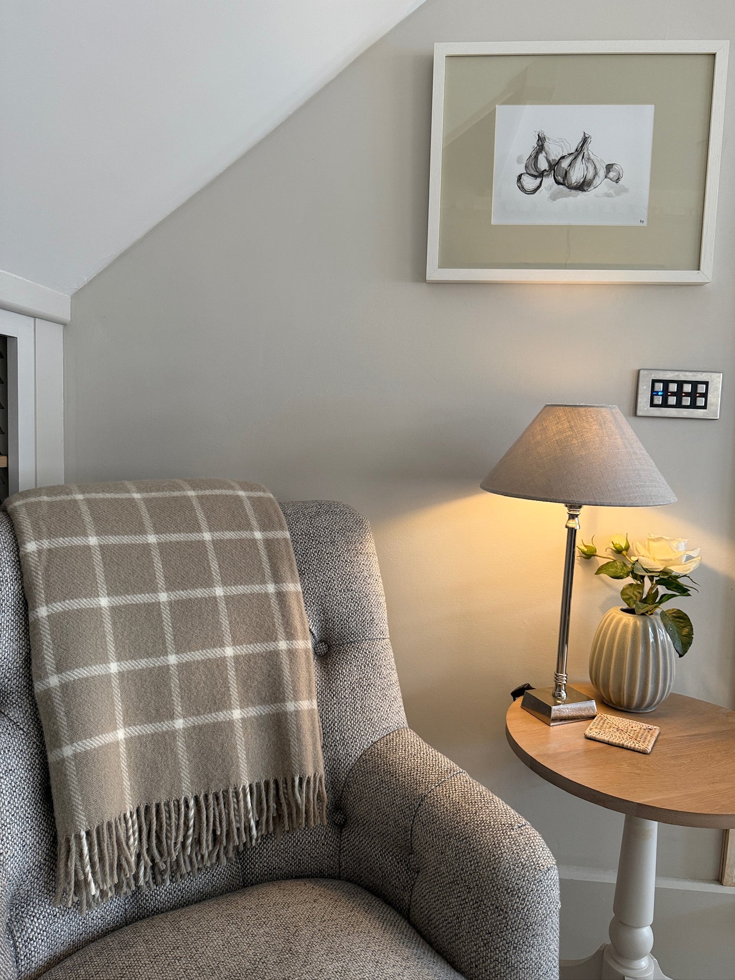 Cozy living room corner with a gray armchair, plaid blanket, lamp, and framed picture.