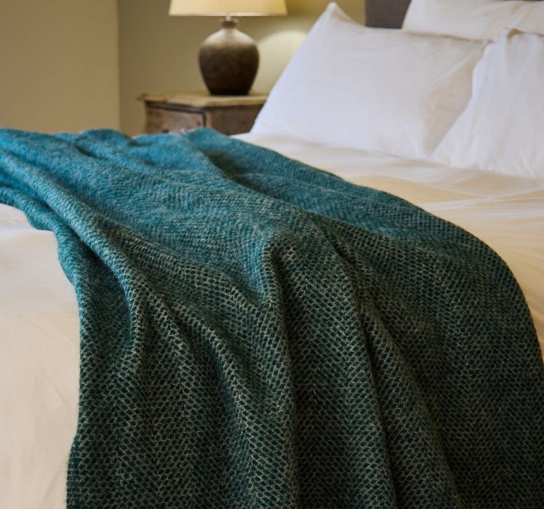 Green textured blanket draped over a bed with white pillows and a lamp in the background.