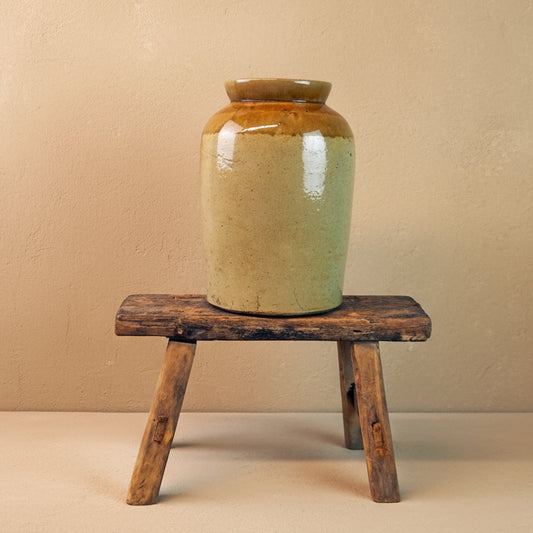 Ceramic jar with a brown top and greenish-brown base on a wooden stool against a plain background