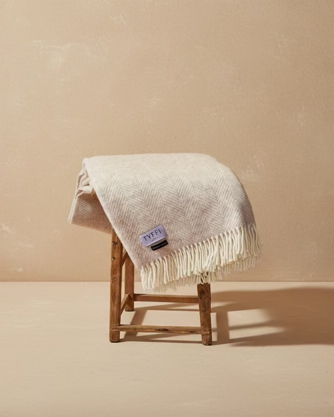 White textured blanket with fringe draped over a wooden stool against a gray wall.