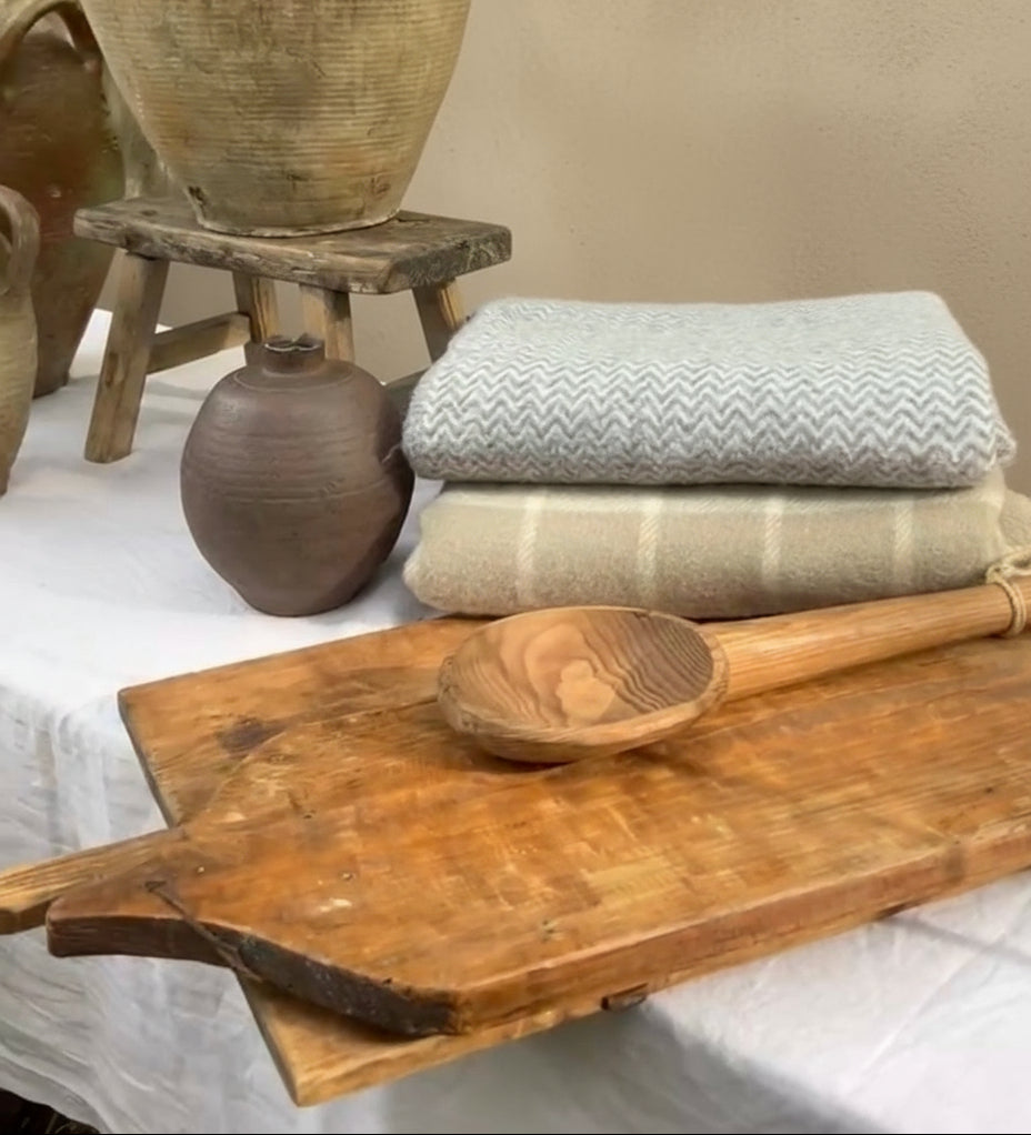 Wooden cutting board with wooden spoon, stacked towels, and ceramic pots on a white surface with greenery in the background.