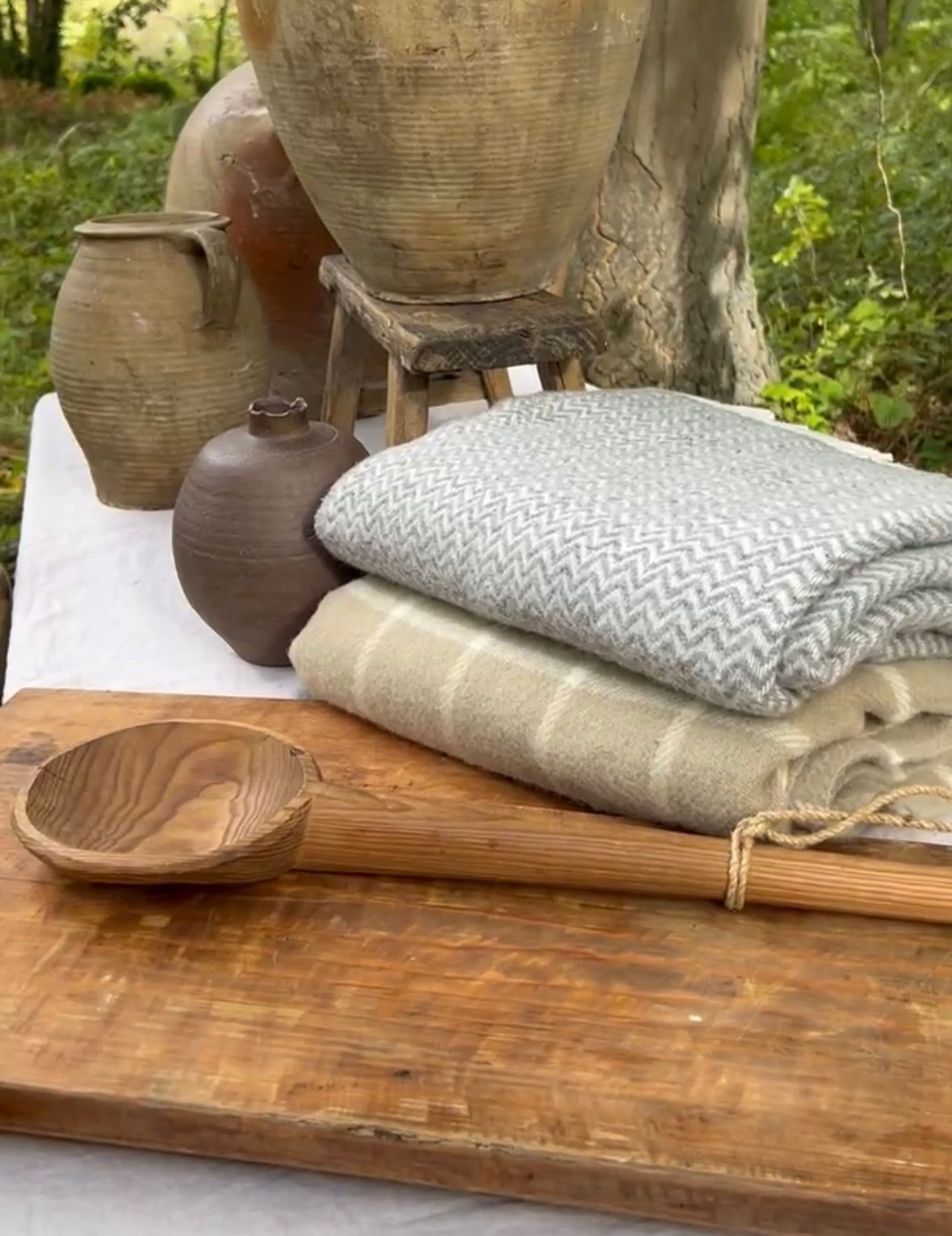 Stack of folded throws on a wooden breadboard with rustic pottery and a stool 