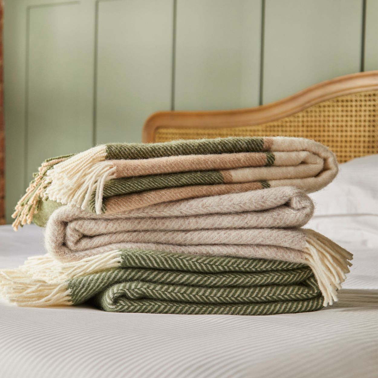 Stack of folded blankets in beige, green, and brown on a bed with a wooden headboard.