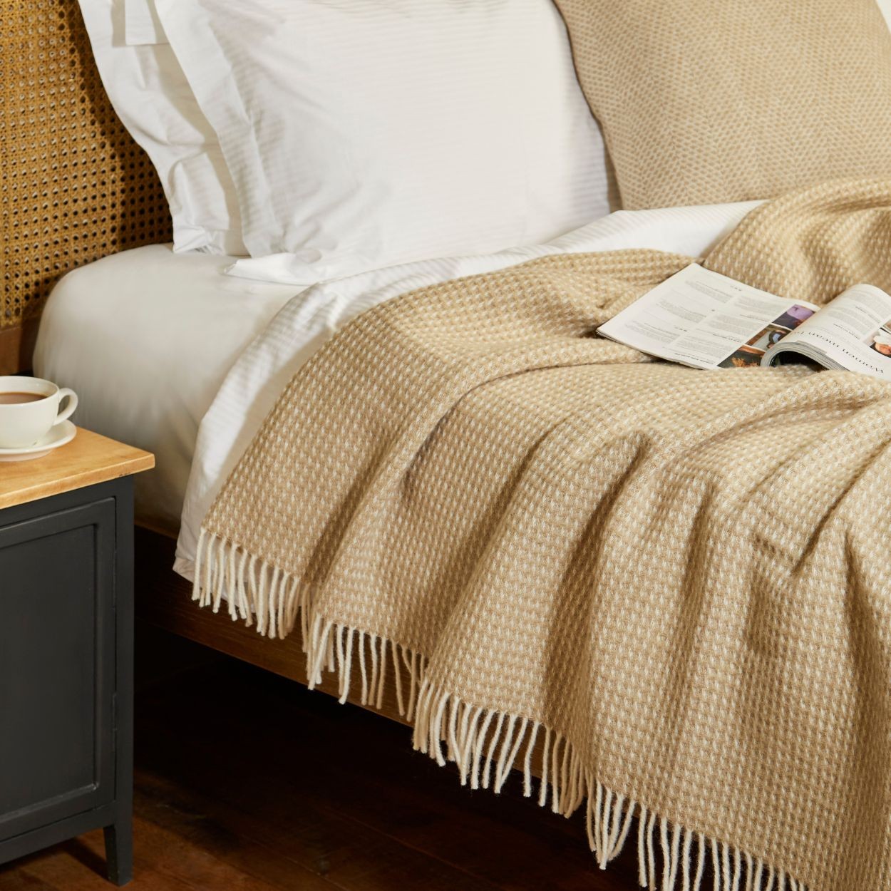 Beige blanket draped over a bed with a side table and newspaper in the foreground